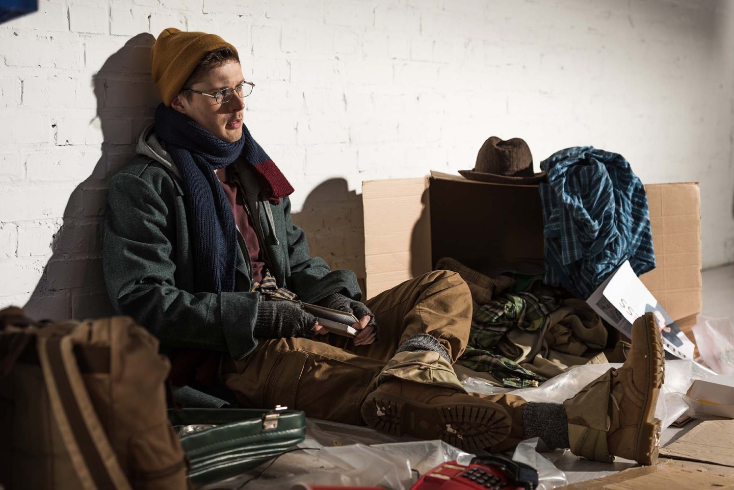 homeless man sitting by brick wall surrounded by rubbish